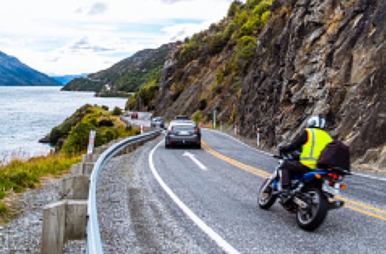 A motorcyclist and some cars going down a narrow lakeside road